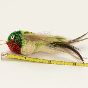 May include: A green, red, and white bird-shaped toy with a long tail of feathers. The toy is about 3 inches long and is measured with a yellow tape measure.