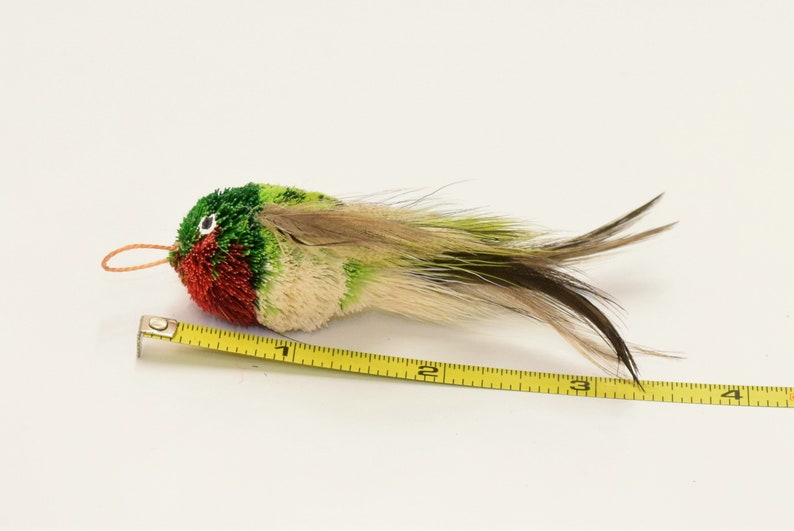 May include: A green, red, and white bird-shaped toy with a long tail of feathers. The toy is about 3 inches long and is measured with a yellow tape measure.