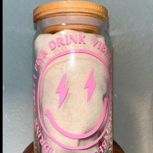 May include: A clear glass reusable drinking cup with a pink smiley face design and the text "Pink Drink Vibes". The cup has a bamboo lid and a clear plastic straw.