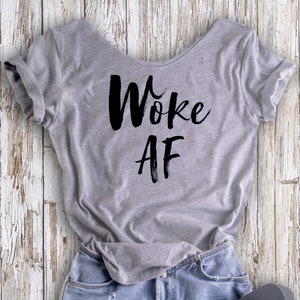 May include: A gray t-shirt with the text "Woke AF" printed in black on the front. The shirt is laid out on a white wooden surface with a pair of blue denim shorts and black flip-flops.