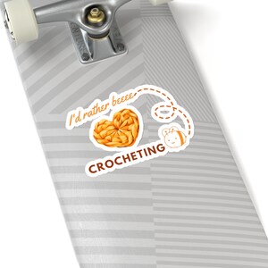 Könnte beinhalten: Ein weißes Skateboard mit einem Aufkleber, auf dem "I'd rather beeee crocheting" steht, mit einem gehäkelten Herzen und einer Bienenillustration.