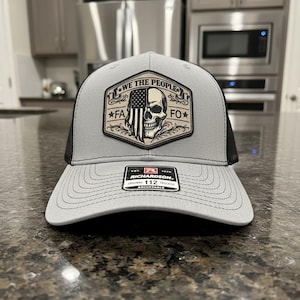 May include: Gray and black trucker hat with a patch featuring a skull, American flag, and the text "WE THE PEOPLE FA FO". The hat has a curved brim and an adjustable snap closure. The Richardson brand label is visible.