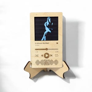 May include: A wooden music player display with a black and blue image of a person. The text "It Almost Worked" and "TV Girl" is visible. The display has a play button, a progress bar, and a QR code.