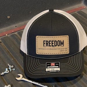 May include: Black and white trucker hat with the word "FREEDOM" on a leather patch. The patch also includes a quote by Charlie Kirk. The hat is a Richardson 112 Original Trucker, adjustable.