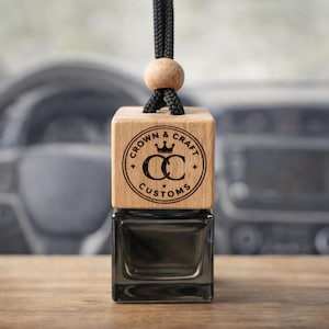 Może przedstawiać: Odświeżacz powietrza do samochodu z drewnianą górą i ciemną szklaną butelką. Drewniana góra jest grawerowana napisem "CROWN & CRAFT CUSTOMS" i logo. Czarny sznurek i drewniana kulka są przymocowane do zawieszenia.
