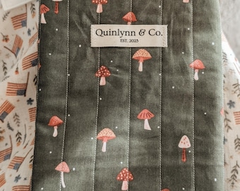 May include: A dark green quilted book cover with a repeating pattern of red and pink mushrooms and white dots. The cover has vertical white stitching and a white ribbon tie. The text "Quinnlynn & Co. EST. 2023" is printed on a white label.