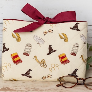 May include: A cream-colored, quilted bag with a burgundy bow, featuring a pattern of wizarding-themed icons. Includes a pair of eyeglasses and a candle in a glass jar. The bag's design includes a sorting hat, owl, and golden snitch.
