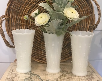 May include: Three white milk glass vases with a scalloped rim and a raised hobnail pattern. The vases are cylindrical with a slightly flared base and top. The center vase contains a bouquet of white flowers and green foliage.