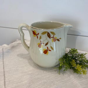 May include: A white ceramic pitcher with a floral design in shades of orange, yellow, and brown. The pitcher has a handle and a spout.