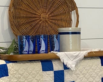 May include: A nautical-themed display featuring blue glasses with sailboat designs, a stoneware crock, and a woven basket. The items rest on a wooden tray atop a blue and white quilt. The basket is round and brown.