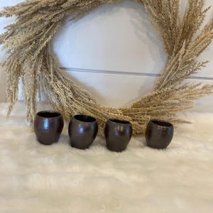 May include: Four small, dark brown, ceramic cups with a textured surface. The cups are arranged in a row on a white, fluffy surface.