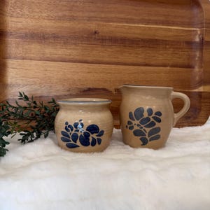 May include: A tan ceramic creamer and sugar bowl set. The creamer has a handle and the sugar bowl has a lid. Both pieces are decorated with a blue floral pattern. The set is on a white fluffy surface with a wooden tray in the background.