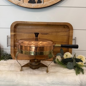 May include: A copper chafing dish with a polished finish, featuring a round lid with a black knob, a long black handle, and a brass-toned metal stand. The base of the stand includes a circular burner component for heating the contents within the pan.
