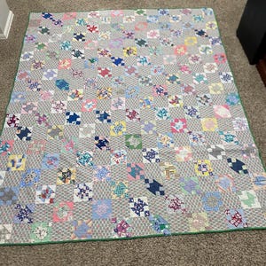 May include: A handmade quilt with a gray background and a patchwork design. The quilt features squares of various colors, including pink, blue, and yellow, with a green border. The quilt is likely made of cotton and is suitable for a bed or as a decorative throw.