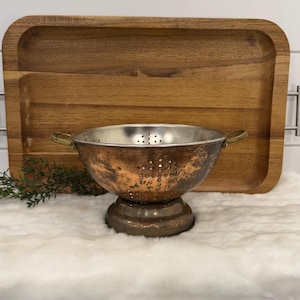 May include: A round metal colander featuring a weathered copper-toned exterior with a patina finish, a silver-toned interior, and two small brass-colored side handles. The bowl is supported by a circular pedestal base and has perforated holes.