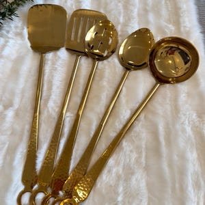 May include: A set of six gold-colored kitchen utensils, including a spatula, slotted spatula, slotted spoon, solid spoon, and ladle. The utensils have long, slender handles and are arranged on a white, fluffy surface. Each handle has a ring at the end.