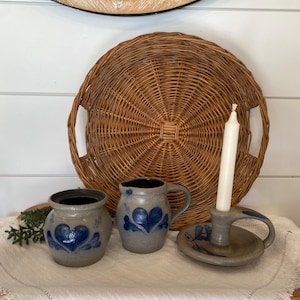 May include: A rustic still life featuring a woven wicker tray, stoneware pottery with blue heart designs, and a candle holder with a lit white candle. The pottery includes a small pitcher and a jar. The background includes a wooden clock.