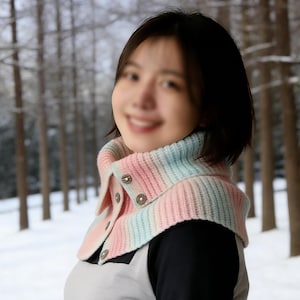 May include: A pastel-colored knitted cowl with a button-up front. The cowl features a gradient of pink, blue, and white stripes. The buttons are silver-toned. The background is a snowy forest.