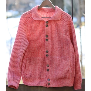 May include: A coral-colored, button-down cardigan sweater with a ribbed knit texture. The sweater has a collared neckline, long sleeves, and a single pocket. The buttons are a dark brown color. The sweater is hanging on a wooden hanger.