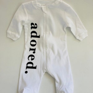 May include: White baby onesie with a black zipper and the word "adored" printed vertically down the front.