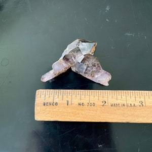 May include: A purple amethyst crystal cluster with a clear, faceted point. The crystal is approximately 2 inches long and is sitting on a wooden ruler.