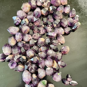 May include: A large group of amethyst crystals, each with a pointed tip, are arranged in a loose pile. The crystals are a deep purple color and have a slightly iridescent sheen.