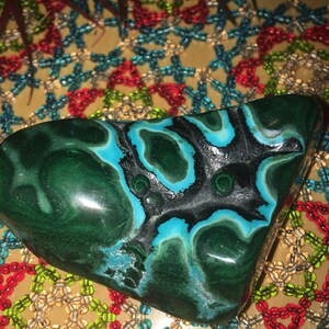 May include: A polished, irregularly shaped stone with a striking pattern of deep green, black, and turquoise blue swirls. The stone's surface is smooth and reflective, showcasing its natural beauty. The stone is a decorative item.