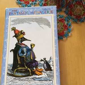 May include: The King of Cups card from The Fantastic Menagerie Tarot deck features a penguin wearing a crown and holding a golden chalice. The penguin sits on a throne made of ice, with a smaller penguin standing behind it. The card has a blue background with a white border.