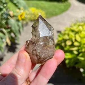 May include: A clear quartz crystal with a smoky brown color and a pointed tip. The crystal is held in a hand.