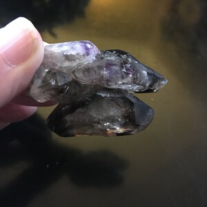 May include: A cluster of raw amethyst and smoky quartz crystals. The amethyst crystals display a vibrant purple hue, while the smoky quartz has a dark, translucent appearance. The crystals are held together, showcasing their natural formations.