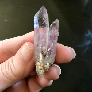 May include: A cluster of two clear quartz crystals with a purple hue. The crystals are pointed and have a natural, rough texture.