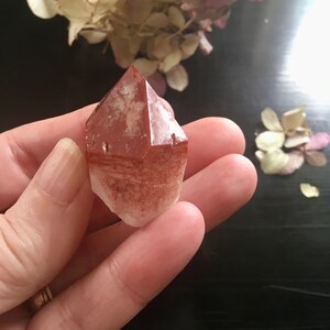 May include: A red and white crystal point with a pointed top and a flat base. The crystal is translucent and has a slight shimmer.