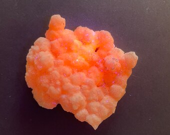May include: A close-up of a cluster of orange-colored crystals with a bubbly texture. The crystals have a gradient of colors, ranging from light orange to a deeper, more vibrant hue. Tiny flecks of purple glitter are scattered throughout.