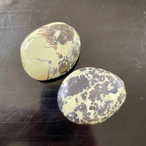 May include: Two oval-shaped stones with a speckled pattern. The stones have a light green base color with dark brown and black markings. The stones are smooth and polished, and they sit on a dark surface.