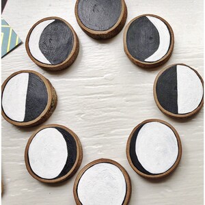 May include: Eight wooden circles with black and white painted moon phases. The circles are arranged in a circle on a white surface.