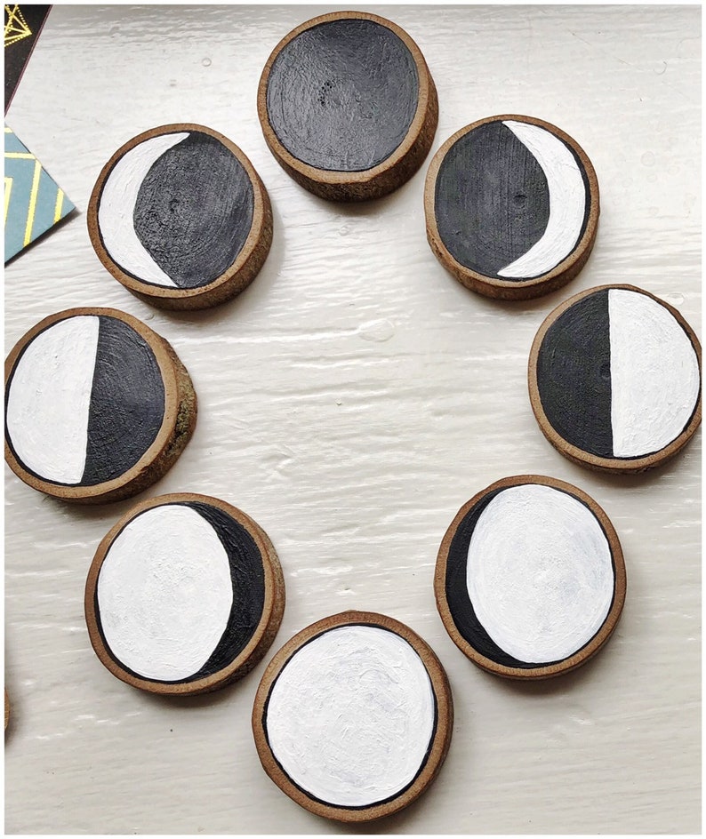 May include: Eight wooden circles with black and white painted moon phases. The circles are arranged in a circle on a white surface.