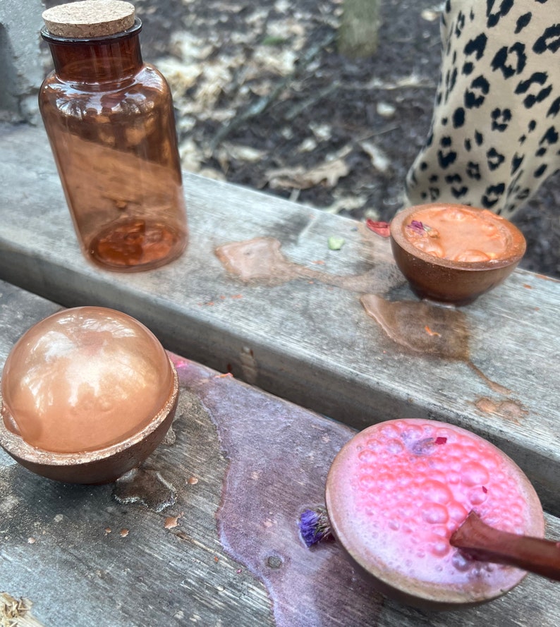 Puede incluir: Tres cuencos peque&ntilde;os llenos de l&iacute;quidos coloridos y burbujeantes. Los cuencos est&aacute;n hechos de madera y tienen un acabado natural. Los l&iacute;quidos son de color rosa, naranja y dorado met&aacute;lico. Los cuencos est&aacute;n sobre una superficie de madera.