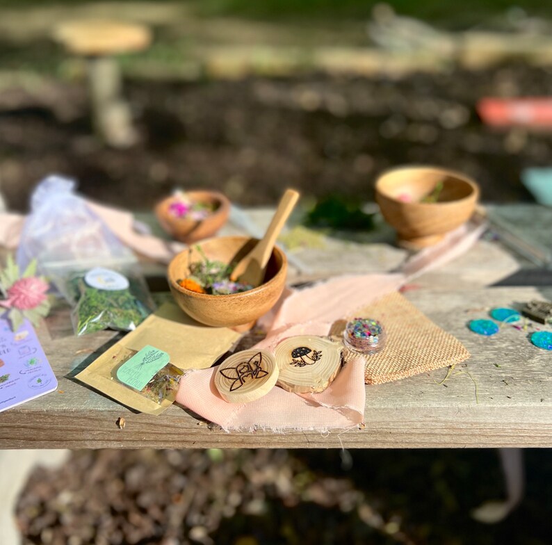 May include: A wooden table with a variety of items, including small bowls, bags of dried herbs, a wooden spoon, and two wooden circles with a butterfly and mushroom design. The table is covered in a light pink cloth and a brown burlap cloth.