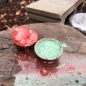 May include: Two resin ornaments, one red and one green, with glitter embedded in the resin. The ornaments are on a wooden surface with glitter scattered around them.