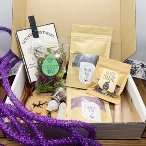 K&ouml;nnte beinhalten: Ein Karton gef&uuml;llt mit verschiedenen Halloween-Artikeln. Enth&auml;lt Beutel mit "Monster Moss" und "Black Magic", einen Totenkopf, einen Holzl&ouml;ffel und eine lila Girlande. Eine Karte mit dem Text "Octopus Tentacles" ist ebenfalls sichtbar.