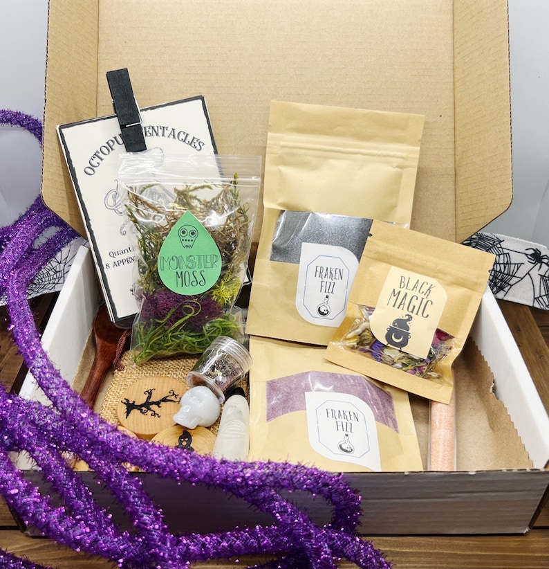 K&ouml;nnte beinhalten: Ein Karton gef&uuml;llt mit verschiedenen Halloween-Artikeln. Enth&auml;lt Beutel mit "Monster Moss" und "Black Magic", einen Totenkopf, einen Holzl&ouml;ffel und eine lila Girlande. Eine Karte mit dem Text "Octopus Tentacles" ist ebenfalls sichtbar.