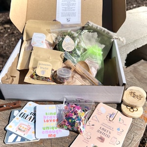 May include: An open box filled with craft supplies for a nature-themed craft project. The box contains dried flowers, moss, glitter, confetti, and cards with positive affirmations. The box also includes a wooden spoon, a glass test tube, and a small wooden disc with a flower design.