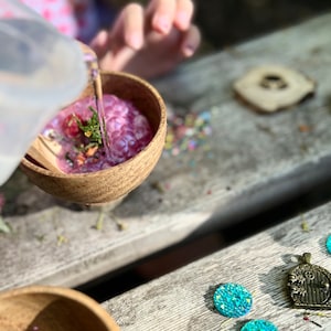 May include: A wooden bowl filled with purple and pink slime, with a clear plastic pitcher pouring water into the bowl. The bowl is sitting on a wooden table with a small wooden circle and blue iridescent gems scattered around it.