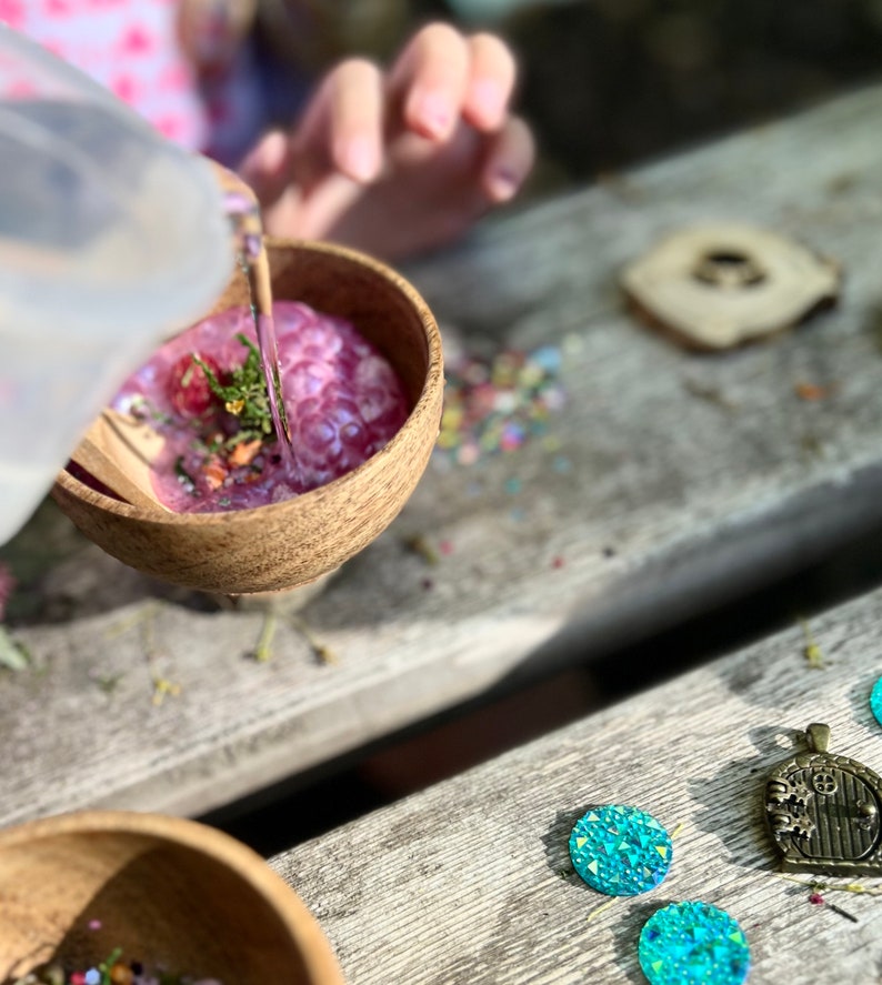 May include: A wooden bowl filled with purple and pink slime, with a clear plastic pitcher pouring water into the bowl. The bowl is sitting on a wooden table with a small wooden circle and blue iridescent gems scattered around it.