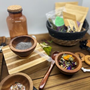 K&ouml;nnte beinhalten: Ein Holztisch zeigt Apothekenartikel. Kleine Holzschalen enthalten Kr&auml;uter, Pulver und getrocknete Blumen. Eine braune Glasflasche mit Korken und ein geflochtener Korb mit Papiert&uuml;ten sind ebenfalls vorhanden.