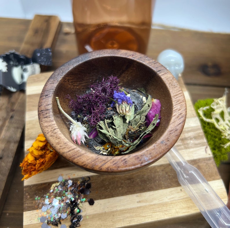 K&ouml;nnte beinhalten: Eine Holzschale gef&uuml;llt mit getrockneten Blumen, Kr&auml;utern und Glitzer. Die Schale steht auf einem Holzschneidebrett mit einer transparenten Pipette und einem Glas Fl&uuml;ssigkeit im Hintergrund. Inhalt: lila, blaue und rosa getrocknete Blumen.