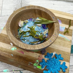 May include: A wooden bowl filled with blue glitter, dried flowers, and a sprig of greenery. The bowl is sitting on a wooden tray with a blue snowflake.