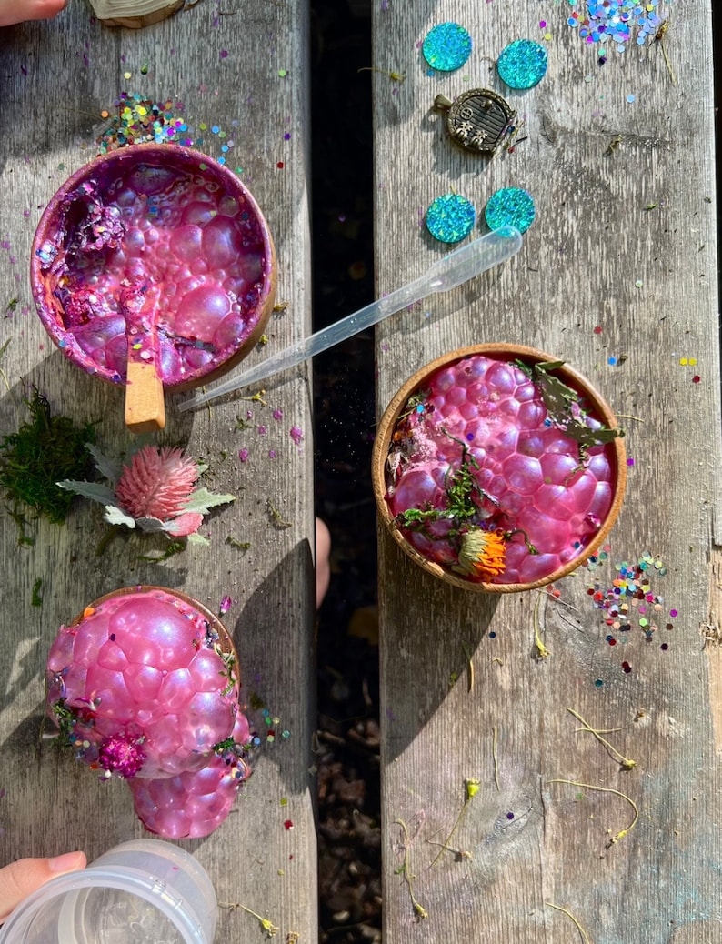 May include: Three bowls filled with pink iridescent slime with glitter and small flowers on a wooden surface.