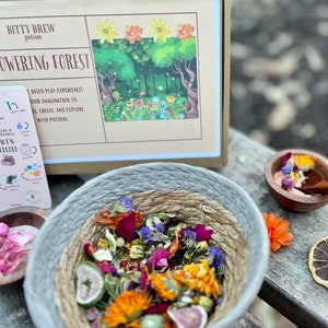 Puede incluir: Primer plano de un cuenco lleno de flores y hierbas secas, etiquetado como "Bitty Brew Potion Flowering Forest". El cuenco est&aacute; sobre una superficie de madera con un peque&ntilde;o cuenco de flores secas y una pieza de fruta en el fondo.