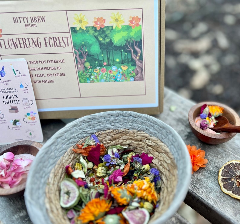 Puede incluir: Primer plano de un cuenco lleno de flores y hierbas secas, etiquetado como "Bitty Brew Potion Flowering Forest". El cuenco est&aacute; sobre una superficie de madera con un peque&ntilde;o cuenco de flores secas y una pieza de fruta en el fondo.